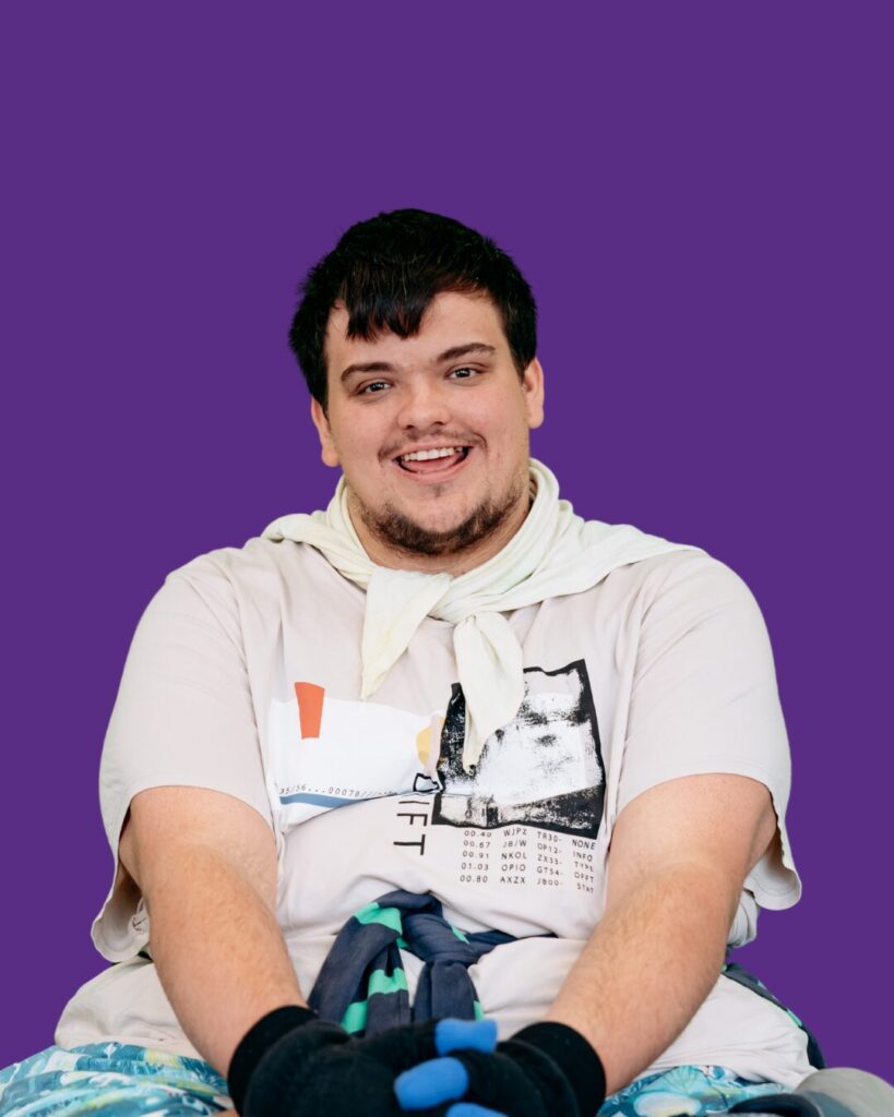 Close-up of participant sitting and smiling. Legs crossed. Cream shirt, blue shorts