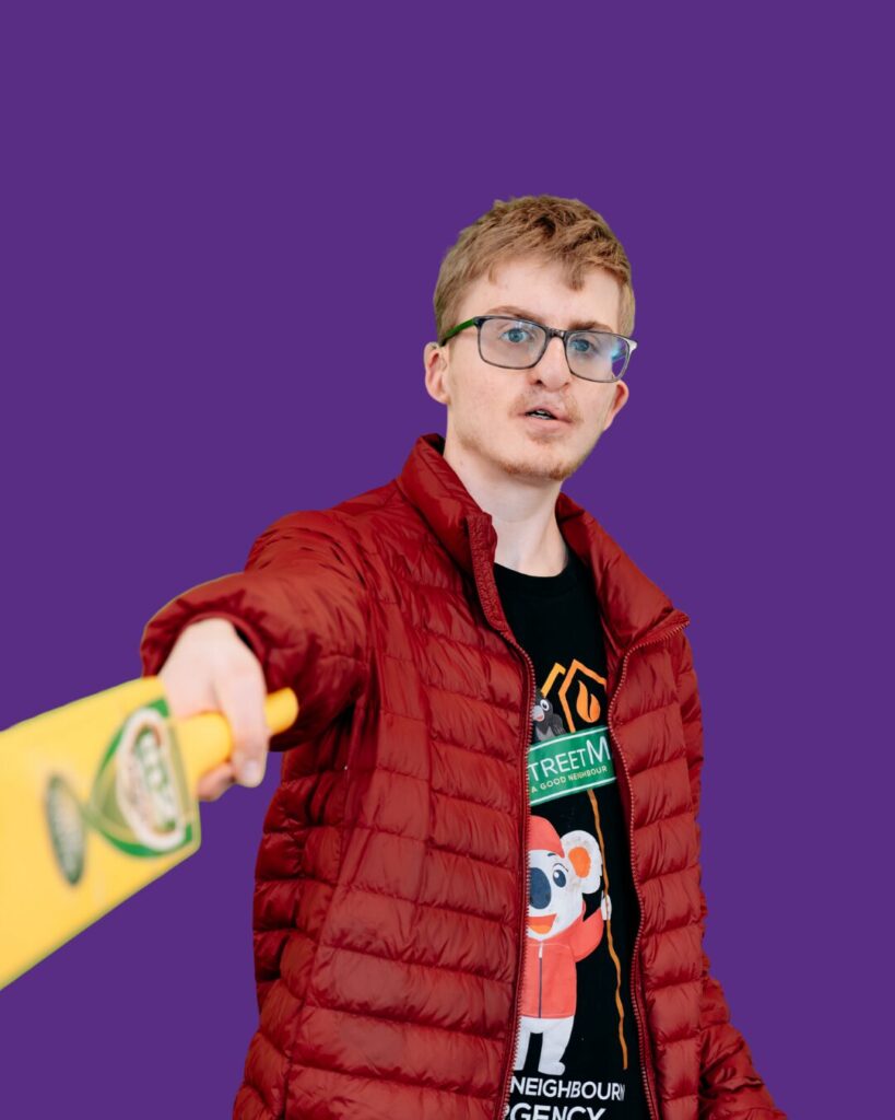 Participant pointing a cricket bat at the camera. Red jack. Ember shirt. Blue jeans